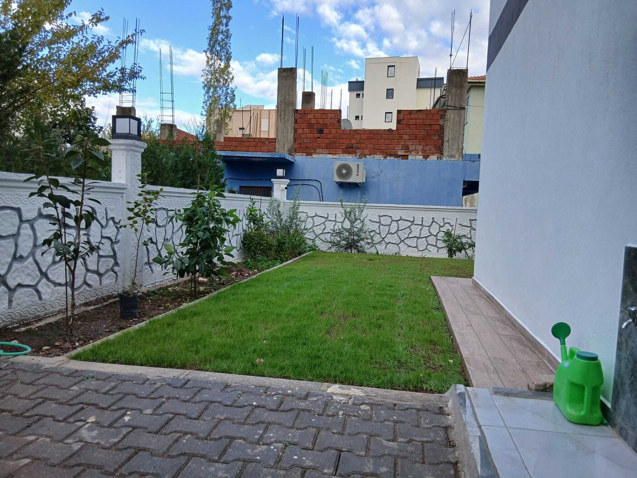 Petit pati pavimentat amb una zona verda, una paret blanca amb treball de pedra decoratiu, i un edifici blau al fons.