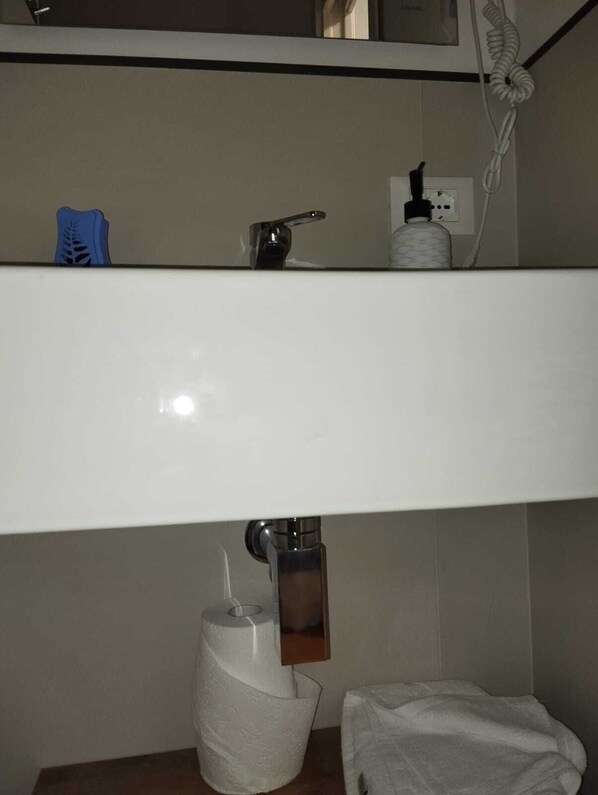 A bathroom with a white sink, a bottle of hand soap, a blue soap dispenser, a toilet paper holder, and a towel on the floor.