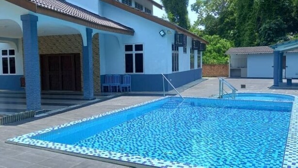 A swimming pool with a tiled deck, a covered area with chairs, and a building with a tiled roof.