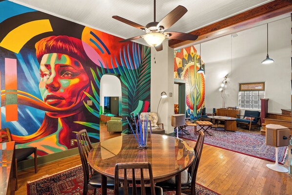 Un coin repas avec une table ronde, des chaises et un ventilateur de plafond. Un mur derrière la table est décoré d’une fresque colorée.