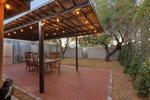 Une terrasse extérieure avec un sol en briques, des chaises en bois et une pergola décorée de guirlandes lumineuses.