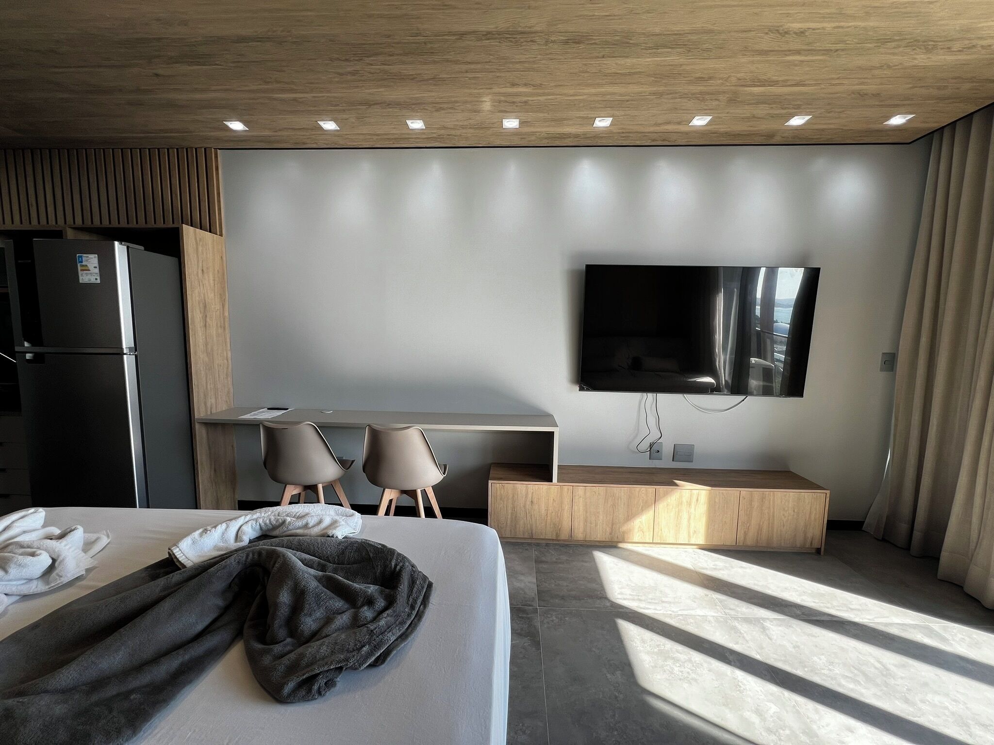 Une chambre d’hôtel moderne équipée d’un lit, d’un bureau, de deux chaises, d’un téléviseur à écran plat et d’un réfrigérateur intégré.
