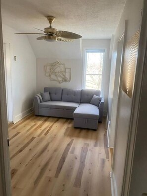 Una habitación con sofá seccional gris, ventilador de techo, piso de madera, una ventana y una obra de arte en la pared.