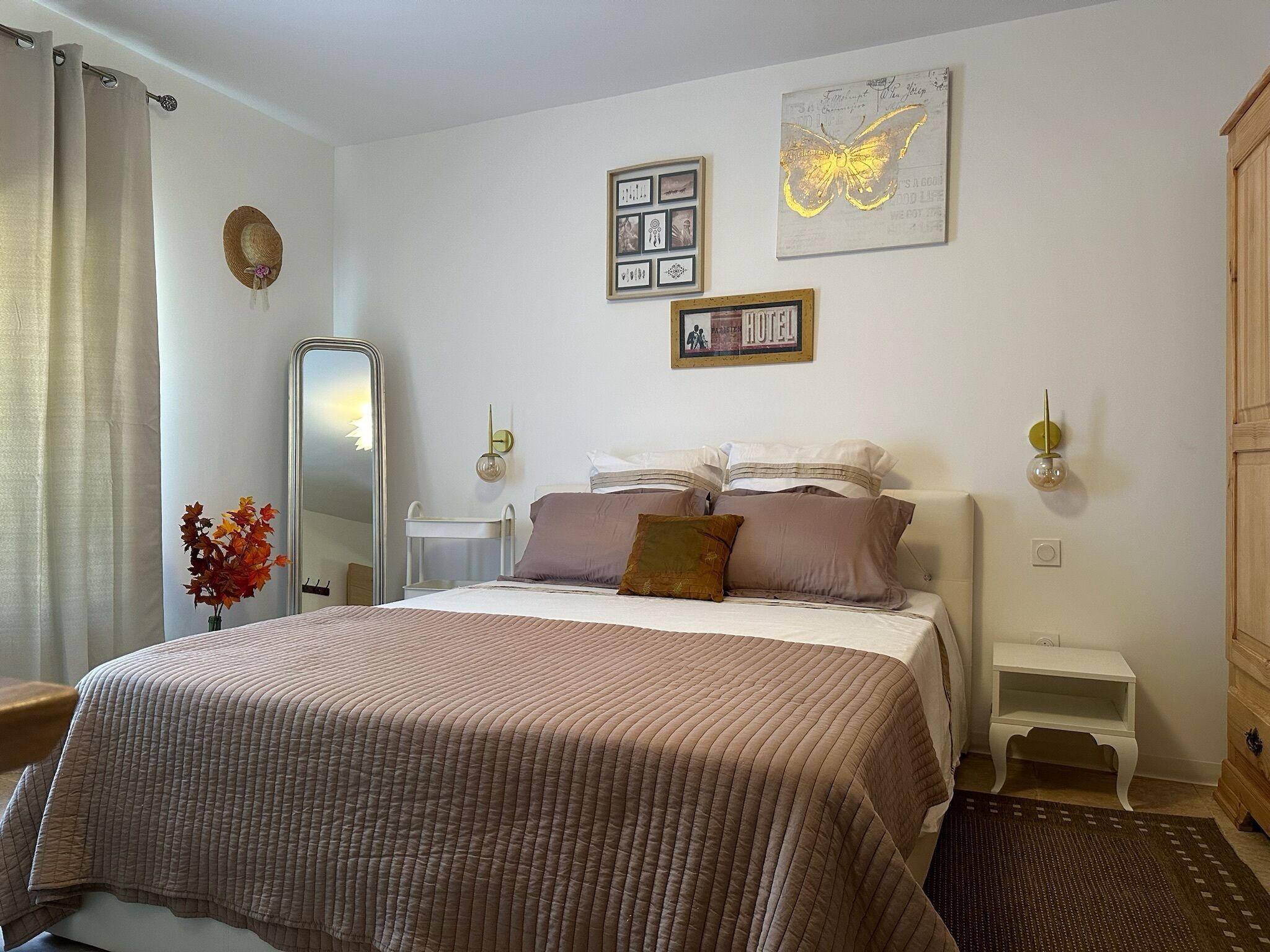 A neatly arranged bedroom with a bed, bedside tables, a mirror, and a wall with framed pictures and a butterfly artwork.