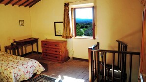 A bedroom with a wooden bed, a dresser, a small table, a window with a view of greenery, and a staircase.