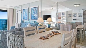 A modern living room with a large window overlooking the ocean, a dining area with a wooden table and chairs, and a cozy seating area with a sofa and cushions. - Fall Deals + Snowbird Rates | Gulf Front Beach Escape (Panama City Beach)