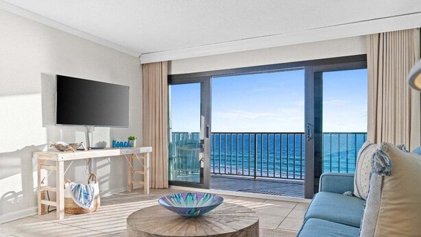 A modern living room with a large TV, a wooden table with a bowl, a sofa, and a balcony with a view of the sea. - Fall Deals + Snowbird Rates | Gulf Front Beach Escape (Panama City Beach)