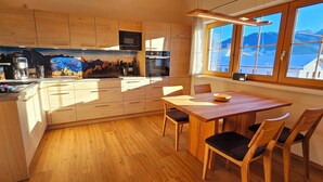 A kitchen with wooden cabinets, a dining table with chairs, and a view of mountains through a window. - Apartment Hipp No. 10 - 67m² new building on the top floor with balcony, mountain and castle view Top location (Halblech)