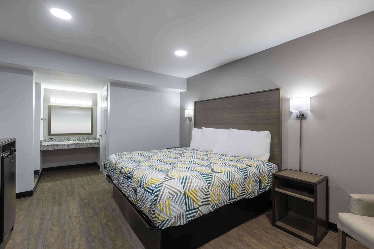 Una habitación de hotel moderna con una cama grande, una mesita de noche, una silla, un escritorio empotrado y un espejo.