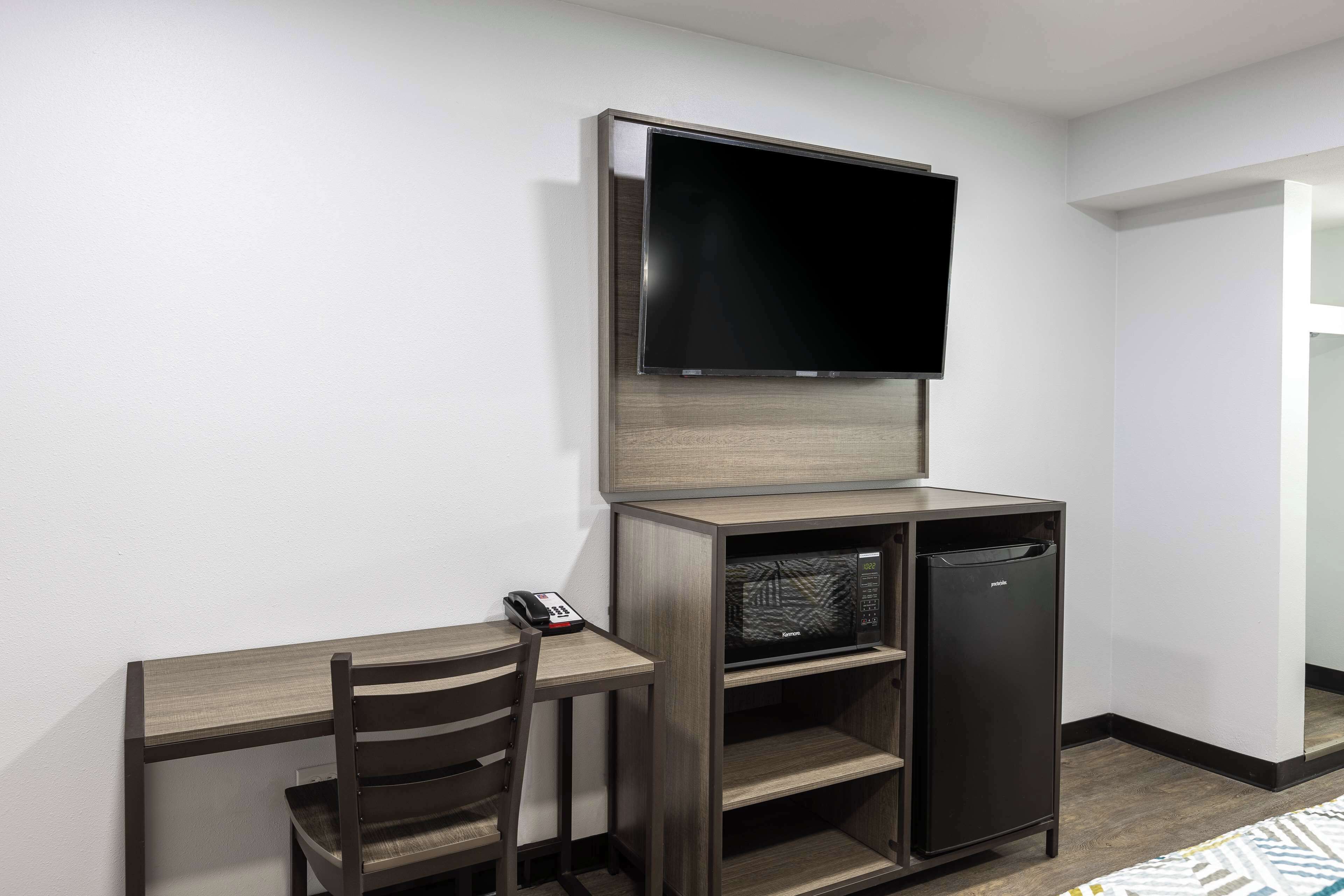Una habitación compacta con una televisión de pantalla plana en la pared, un escritorio de madera pequeño con una silla, y un microondas integrado debajo de la televisión.