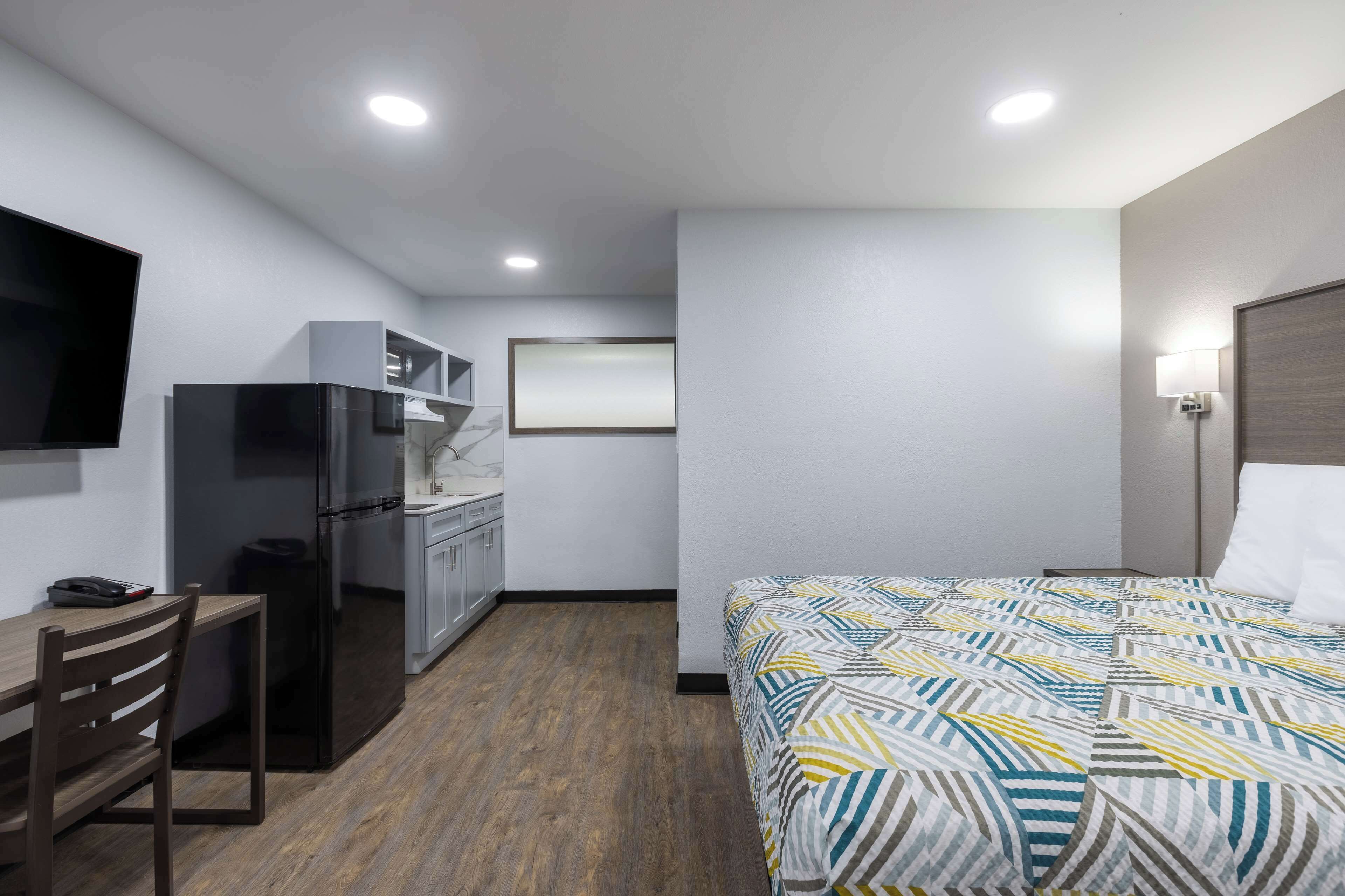 A compact hotel room with a bed, a desk, a kitchenette, and a TV.