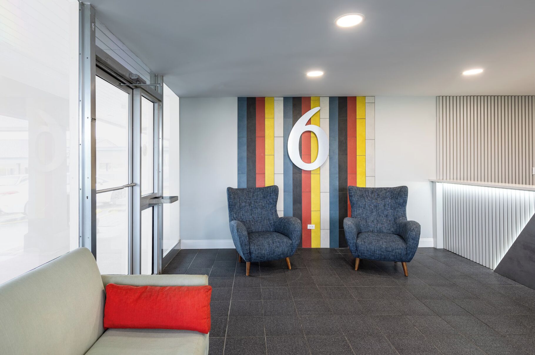 Een moderne wachtruimte met twee blauwe fauteuils, een rood kussen op een grijze bank, en een kleurrijke gestreepte muur met het grote getal '6'.