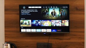 Uma televisão fixada numa parede de madeira exibindo uma interface de streaming com diversas opções de conteúdo.
