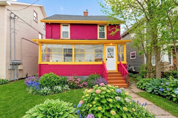 A two-story house with a red exterior and yellow trim, a front porch with steps, and a garden with various plants.