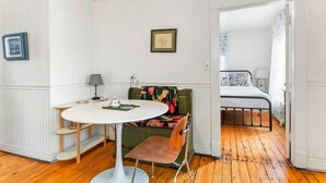 Una habitación con una mesa redonda, un banco con tapicería floral, una cama con colcha estampada y un estante pequeño con una lámpara.