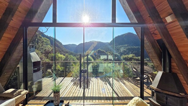 A modern loft with large glass windows, a wooden deck, and a view of a mountainous landscape. - Cabanas Lumberjack (Urubici)
