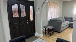 Uma sala de estar com uma porta de madeira escura, um sofá cinza, uma mesinha e um tapete estampado.