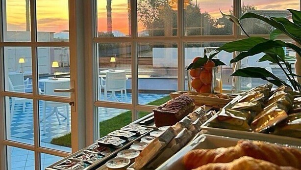 A breakfast buffet with pastries and sandwiches, a pool area visible through large windows, and a view of palm trees and a sunset. - Villa Oteri Room & Bistrot (Bacoli)