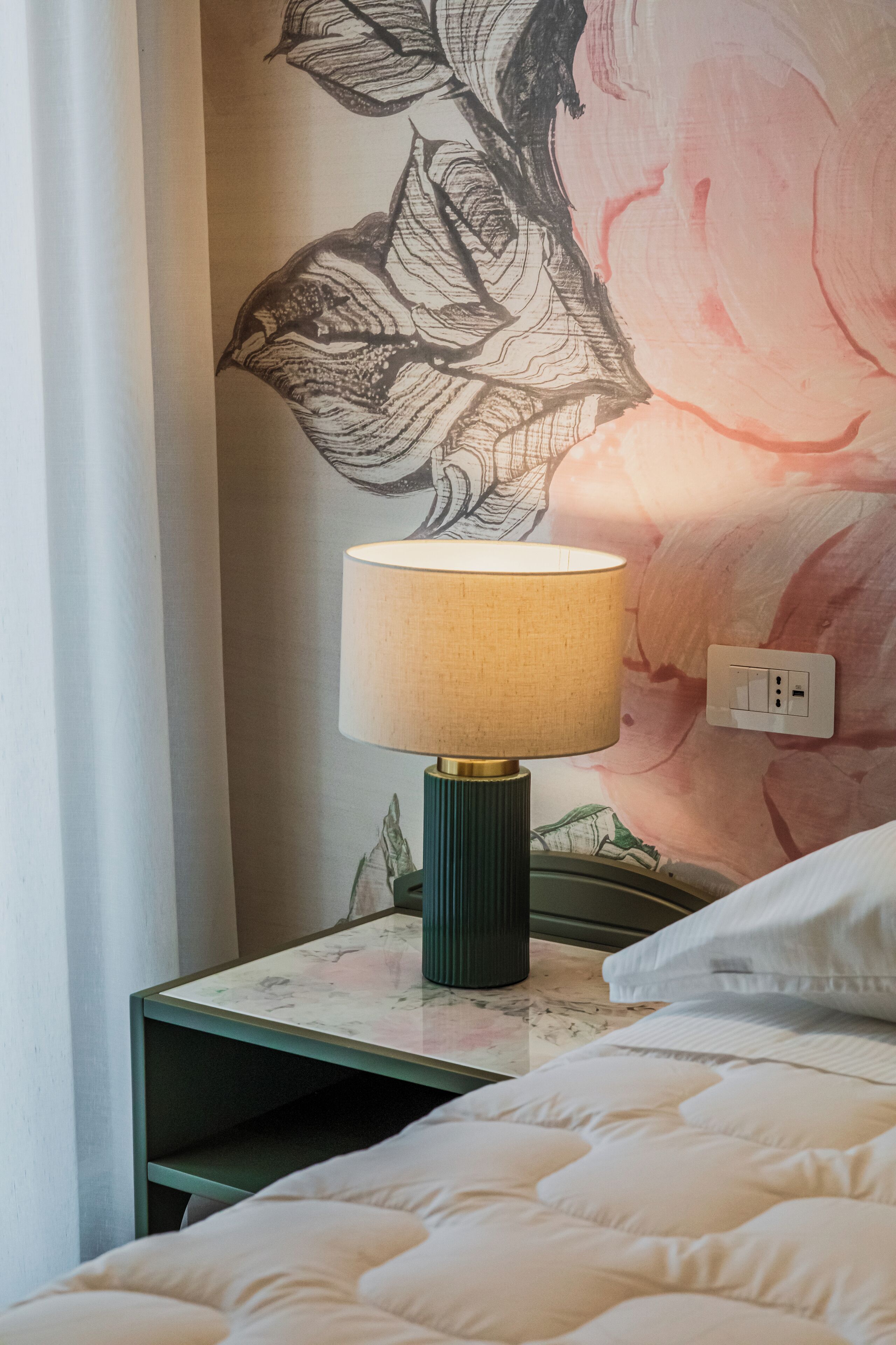Una cama con edredón blanco, una mesita de noche verde con lámpara y una pared con un mural floral.