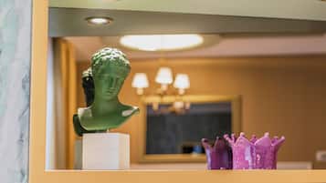 Pared de mármol con un espejo enmarco que refleja un busto de escultura verde, una corona púrpura y una placa blanca con texto.