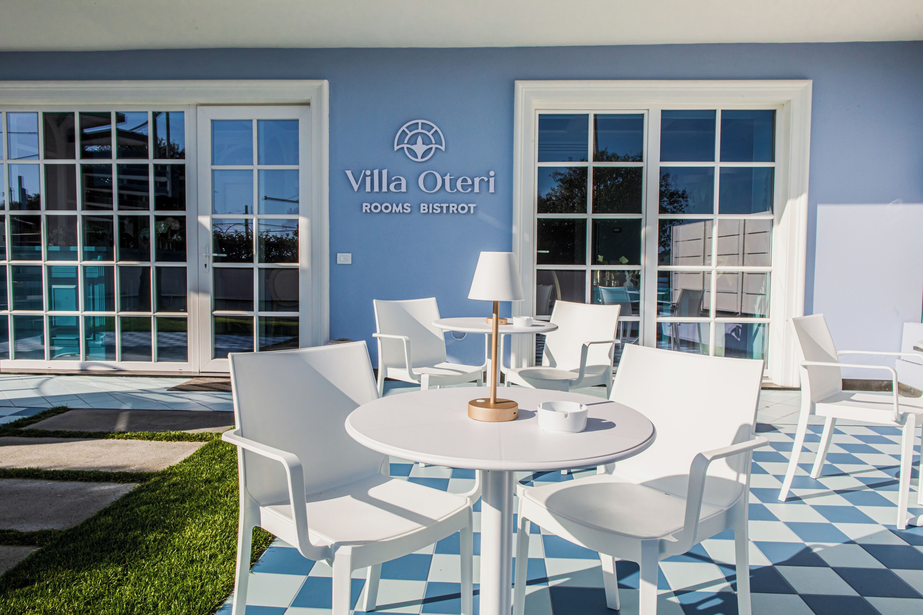 Et udendørs siddeområde med hvide stole og borde, et skilt med teksten "Villa Oteri Rooms Bistro" og et skakternet gulvmønster.