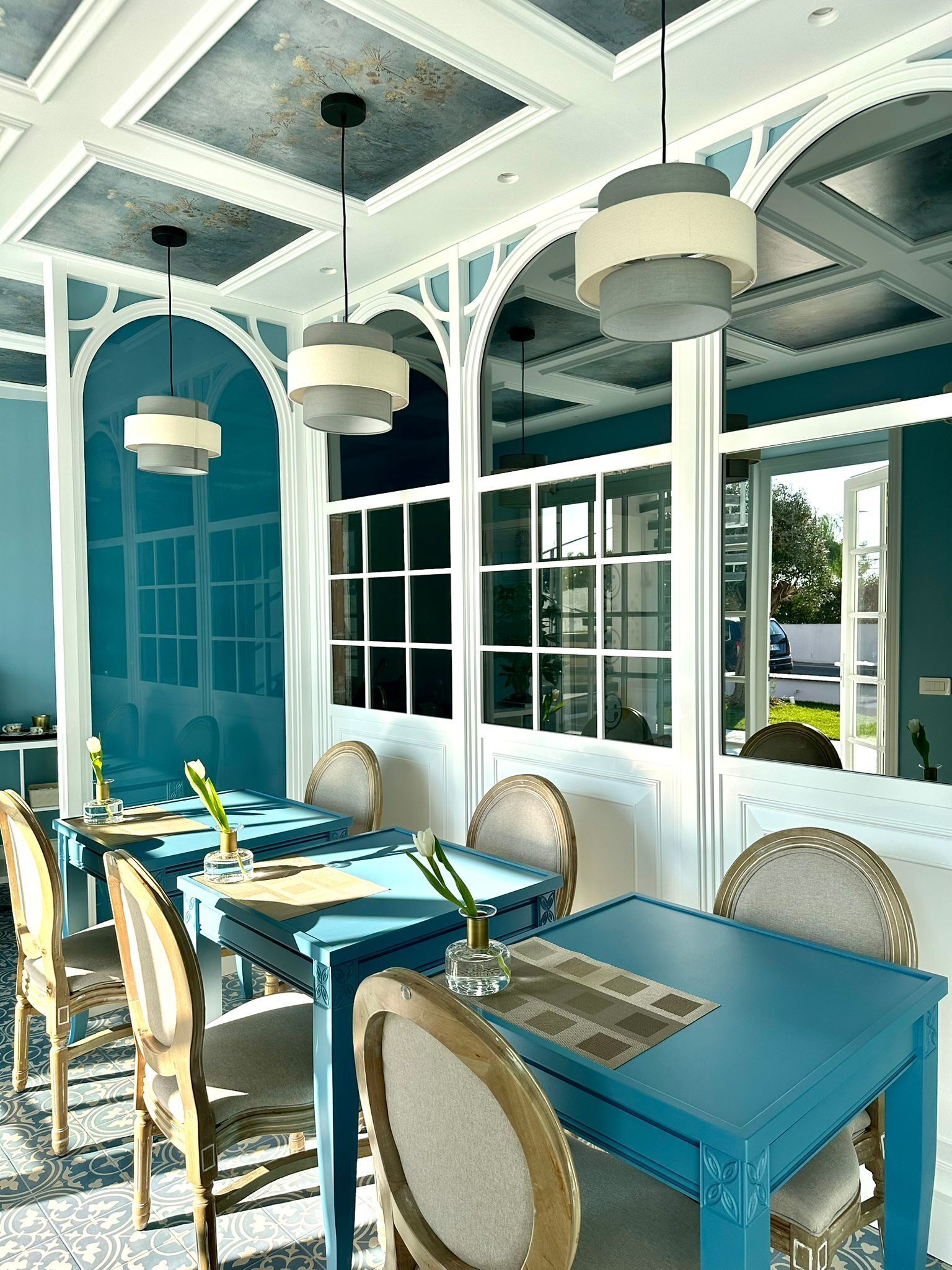 A dining area with blue tables and chairs, large windows, and hanging pendant lights.