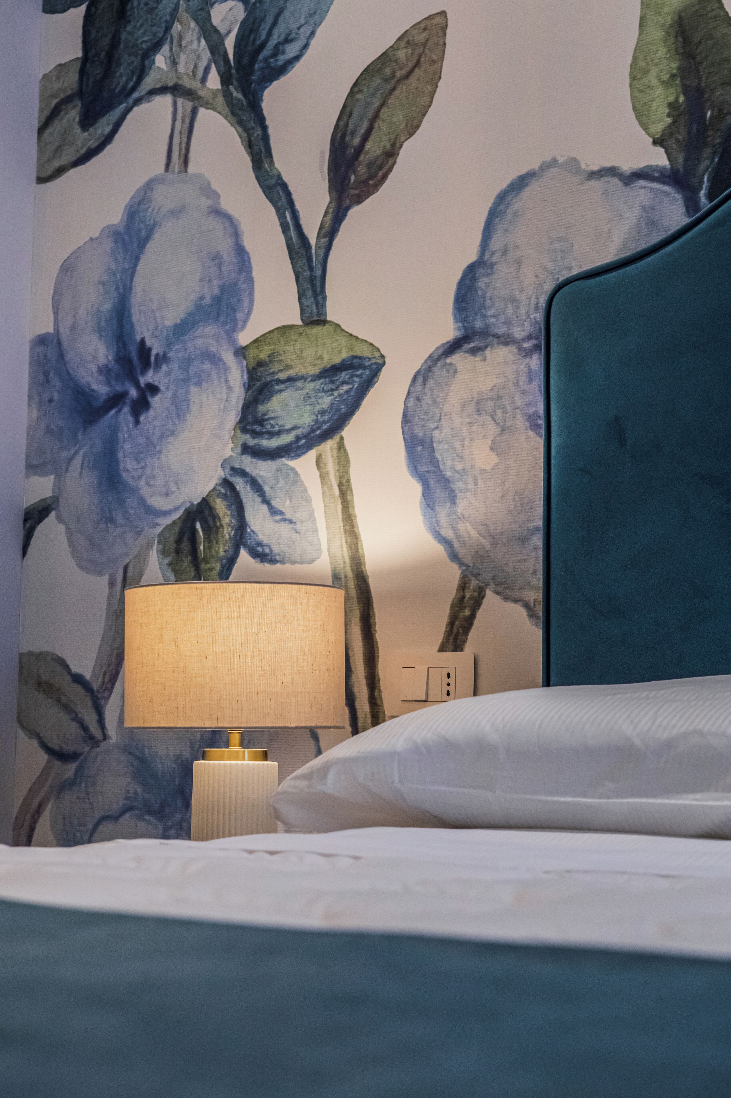 Una habitación de hotel con una cama, un cabecero azul, una pared con papel tapiz floral, una lámpara de noche y una mesita de noche.