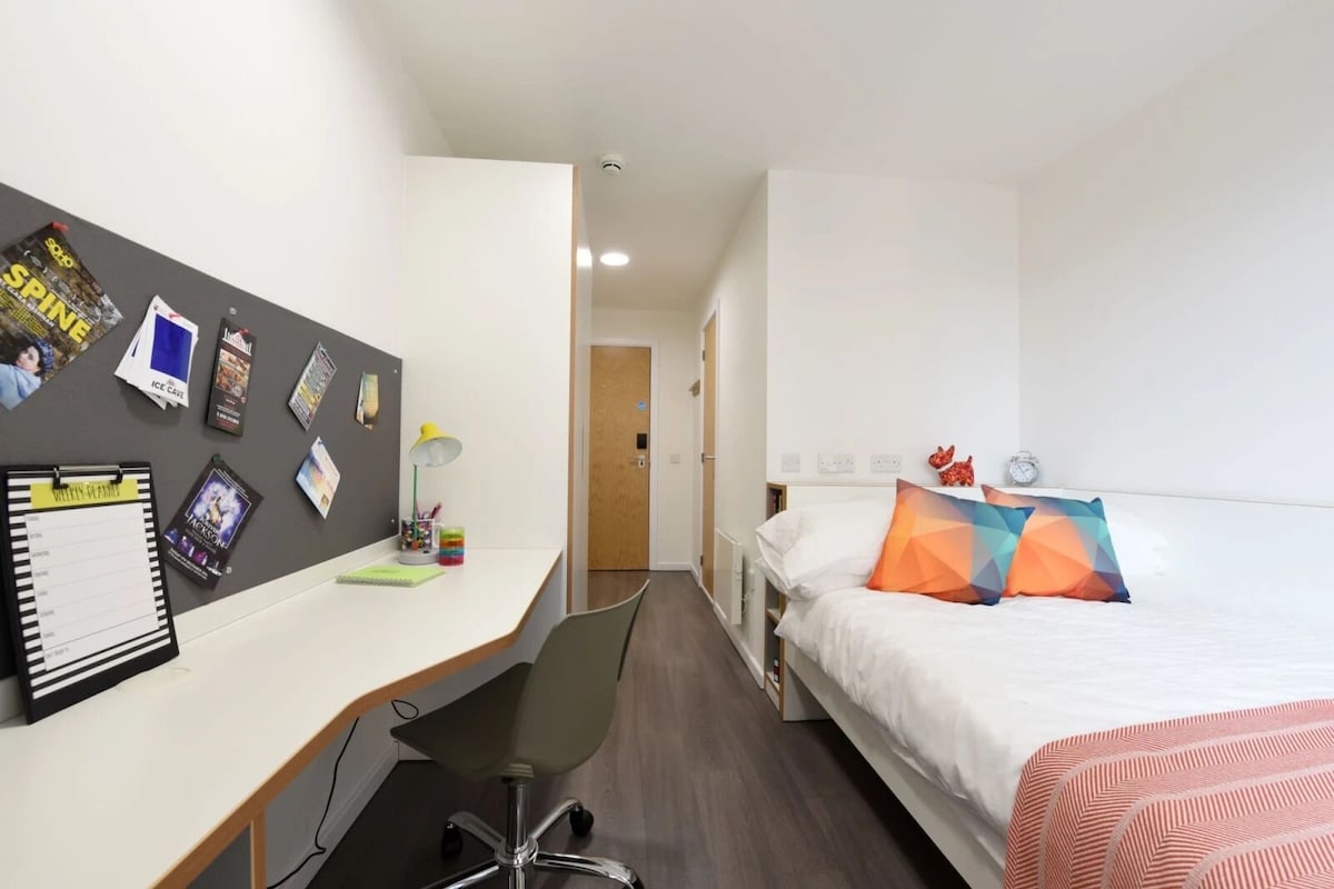 A hotel room with a desk, chair, bed, and a wall with various items pinned to it.