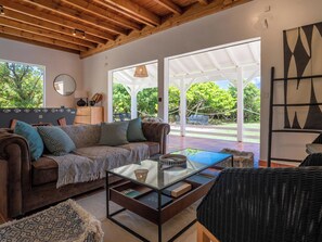 Un salon chaleureux avec un plafond en bois, un canapé agrémenté de coussins à motifs, une table basse en verre et une vue sur l’extérieur grâce à de grandes fenêtres.