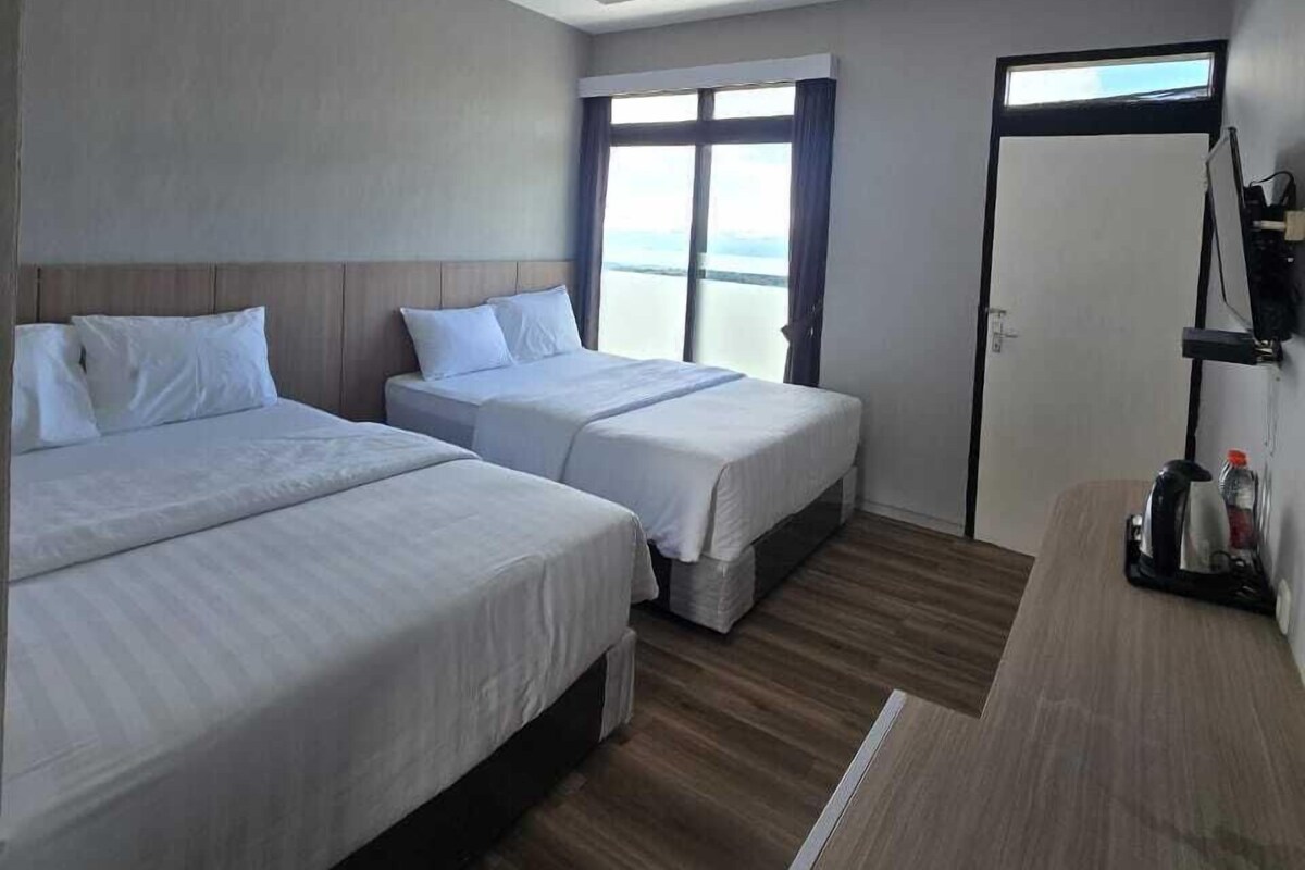 Habitación de hotel con dos camas individuales, piso de madera, un ventanal grande con cortinas y una zona de kitchenette.