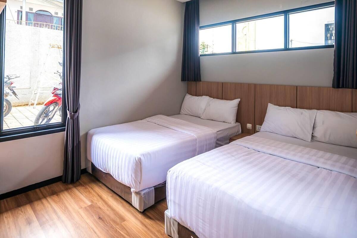 A hotel room with two beds, wooden flooring, a window with curtains, and a view of the outside.
