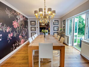 A dining area with a long wooden table, white chairs, a chandelier, framed pictures on the wall, and a view of the outdoors through large windows.