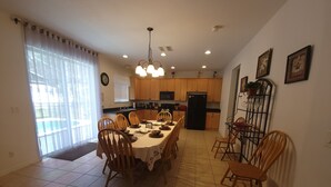 A dining area with a large table set for six, surrounded by wooden chairs. There is a kitchen area with wooden cabinets and a black refrigerator.