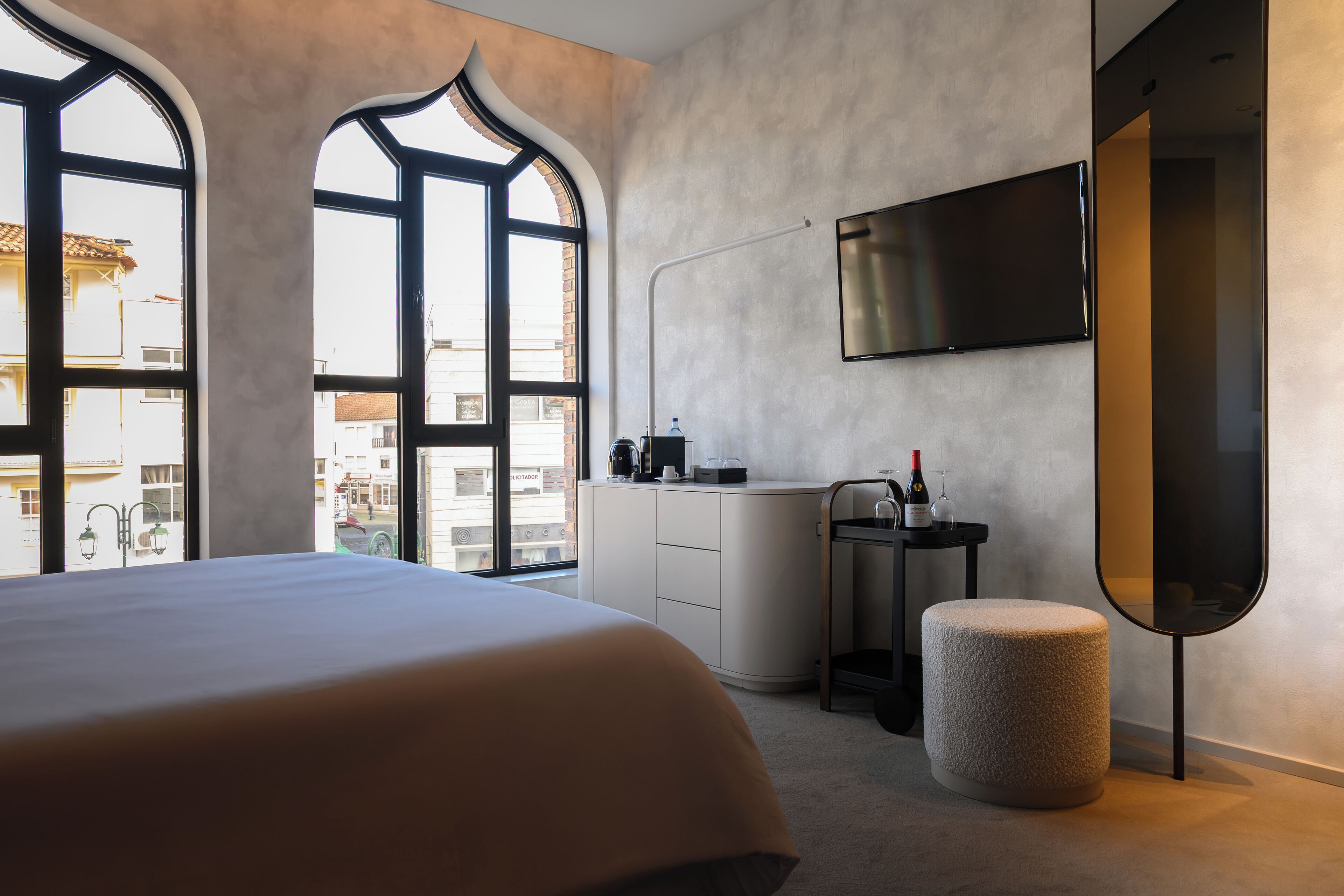 A modern hotel room with a large bed, a TV mounted on the wall, a small table with a bottle and glasses, and a view through large arched windows.