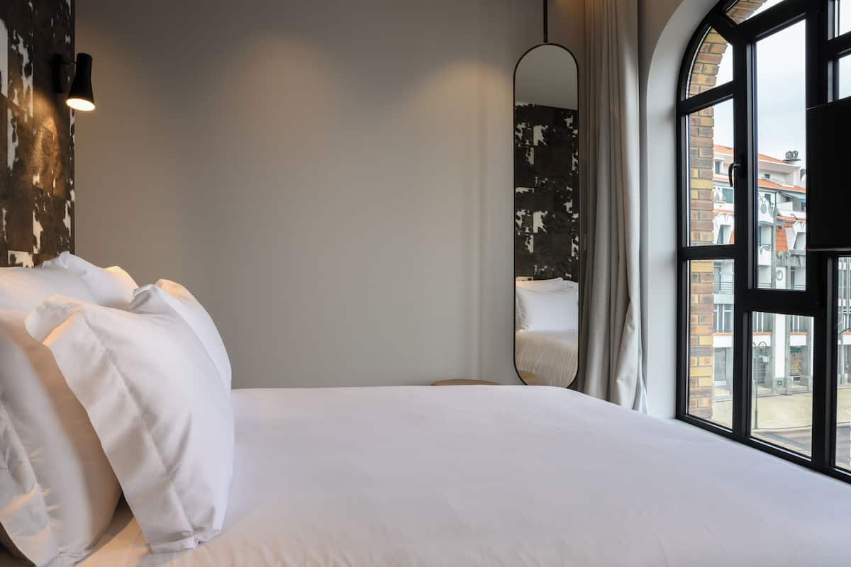 A hotel room with a large bed, a mirror, and a view of buildings through a window.