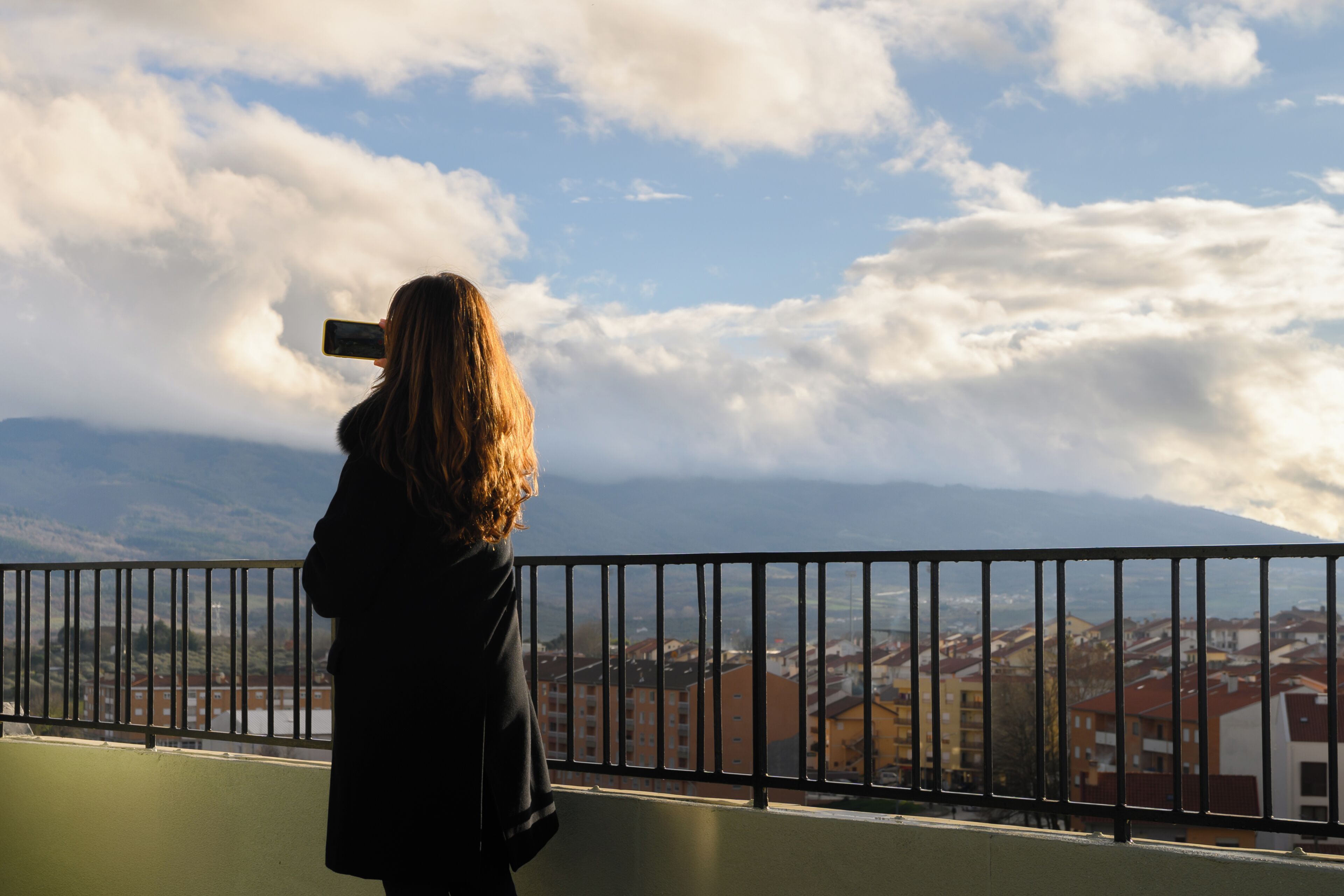 Una persona tomando una foto con un teléfono inteligente, con vistas a un paisaje urbano que incluye edificios y montañas al fondo.