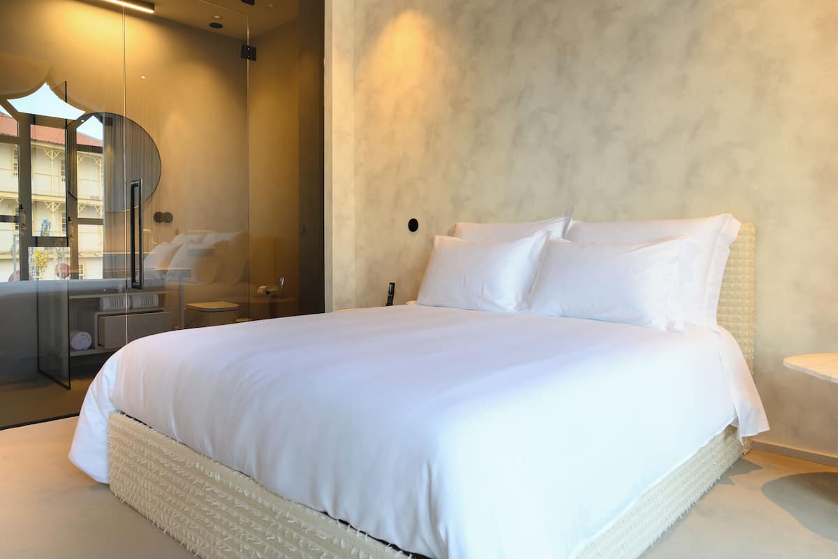A modern hotel room with a large bed, a bedside table, a mirror, and a view of the outside.