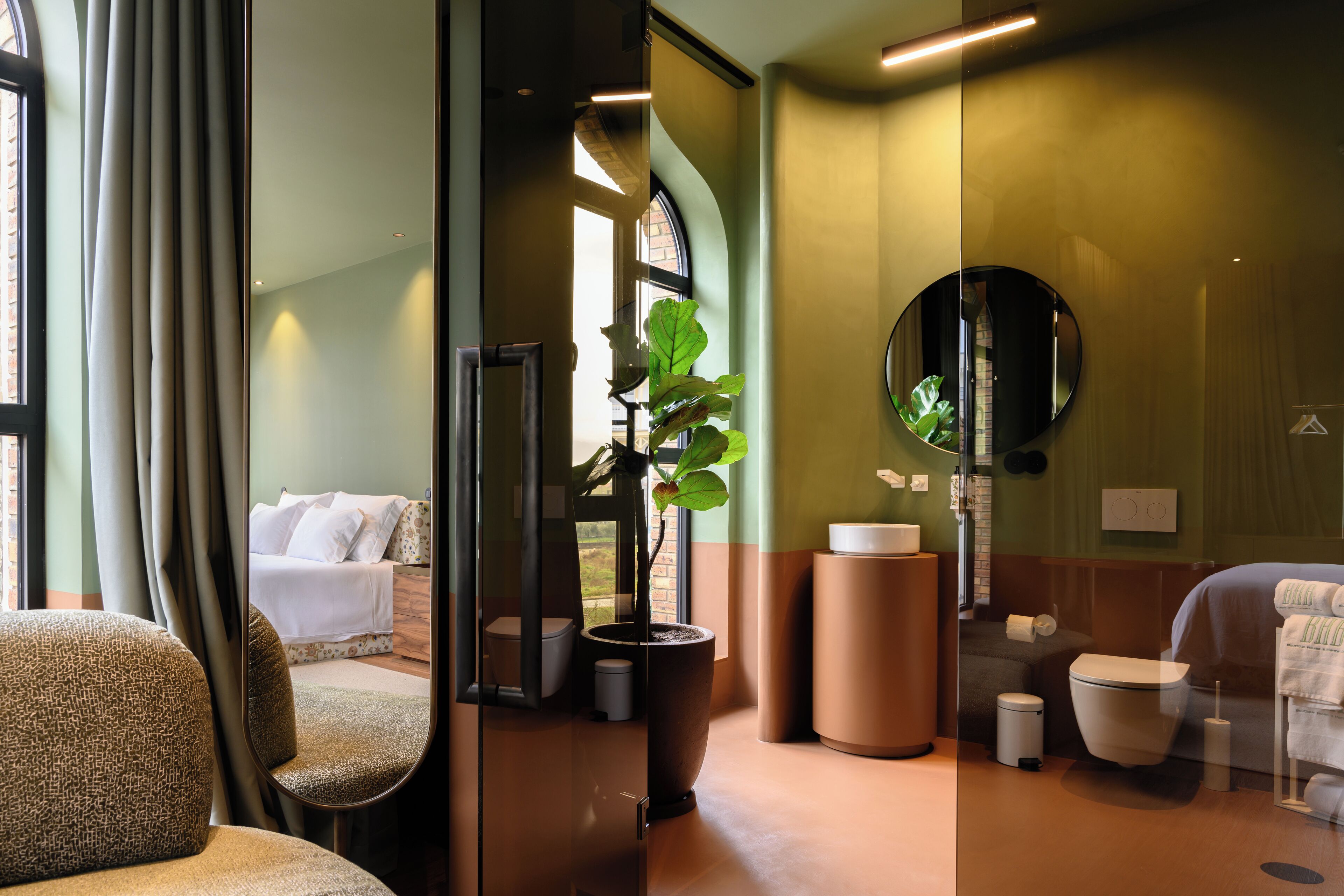 Un dormitorio moderno con un espejo grande, una planta en maceta, una cama, un baño con un espejo redondo y vistas al exterior.