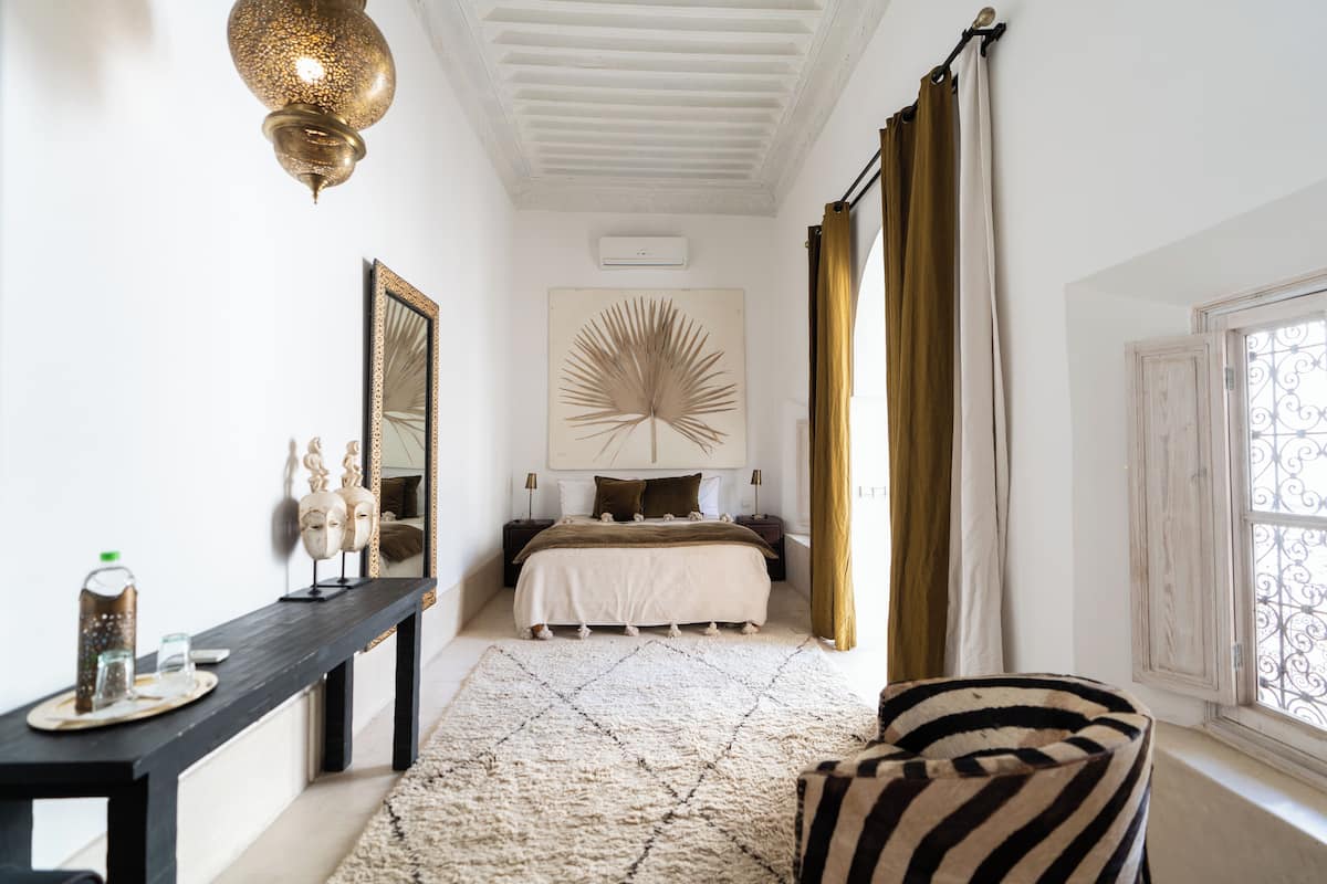 A bedroom with a bed, a nightstand, a mirror, a wall-mounted fan, and a window with curtains.