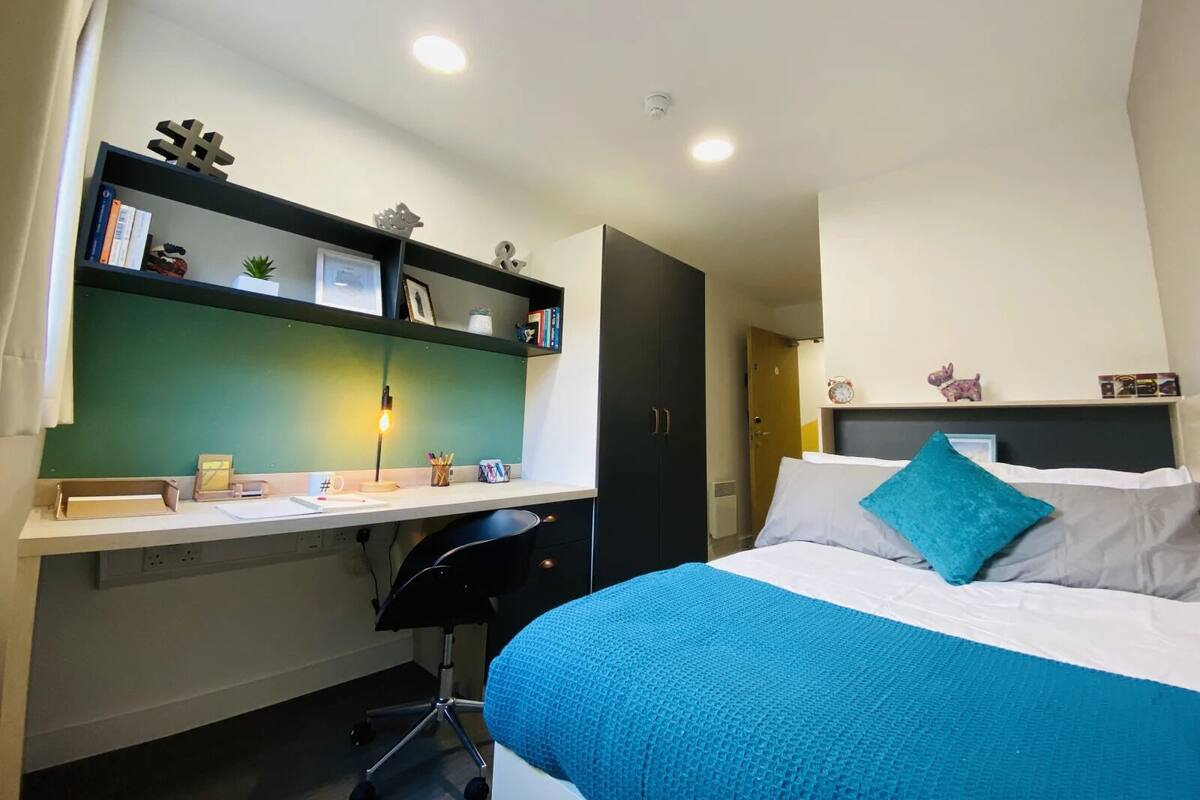 A bedroom with a bed, a desk, and a chair. There is a shelf with books and decorative items.