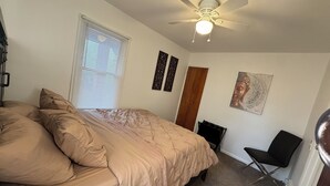 Un dormitorio con cama, ventilador de techo, ventana con persianas, puerta y una silla.