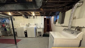 Lavandería con lavadora, secadora y lavabo. Se ve una escalera de madera y un estante con productos de limpieza.