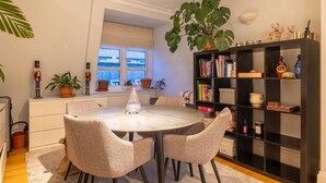 Ein Essbereich mit einem runden Tisch, vier Stühlen, einem großen Topfpflanzen, einem Bücherregal und einem Fenster mit Jalousien.