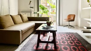 A modern living room with a beige sofa, a glass coffee table, a patterned rug, a brown leather chair, and a view of greenery outside.