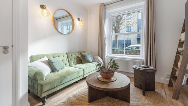 Un salon avec un canapé vert, une table basse ronde ornée d’une plante en pot, une petite table d’appoint ronde, un grand miroir et une fenêtre avec des rideaux.