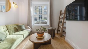 Un salon avec un canapé vert, une table basse ronde sur laquelle est posée une plante en pot, une fenêtre donnant sur des immeubles et une échelle en bois appuyée contre le mur.
