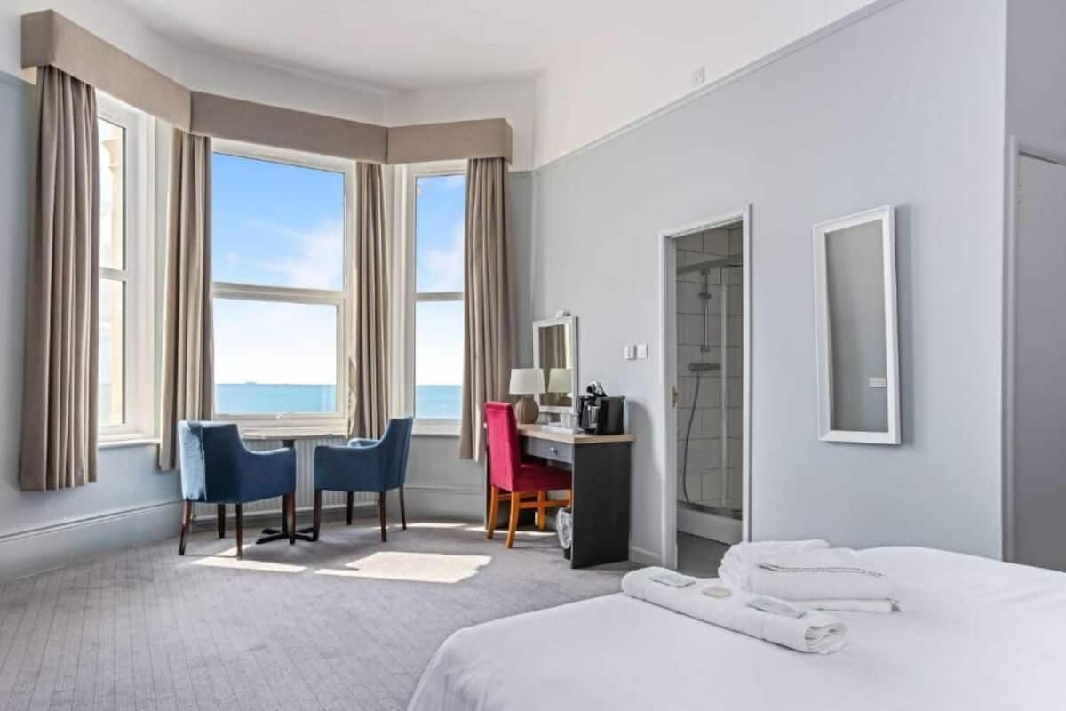 Habitación de hotel con cama, escritorio, dos sillas, lámpara de escritorio, espejo y vista al mar.