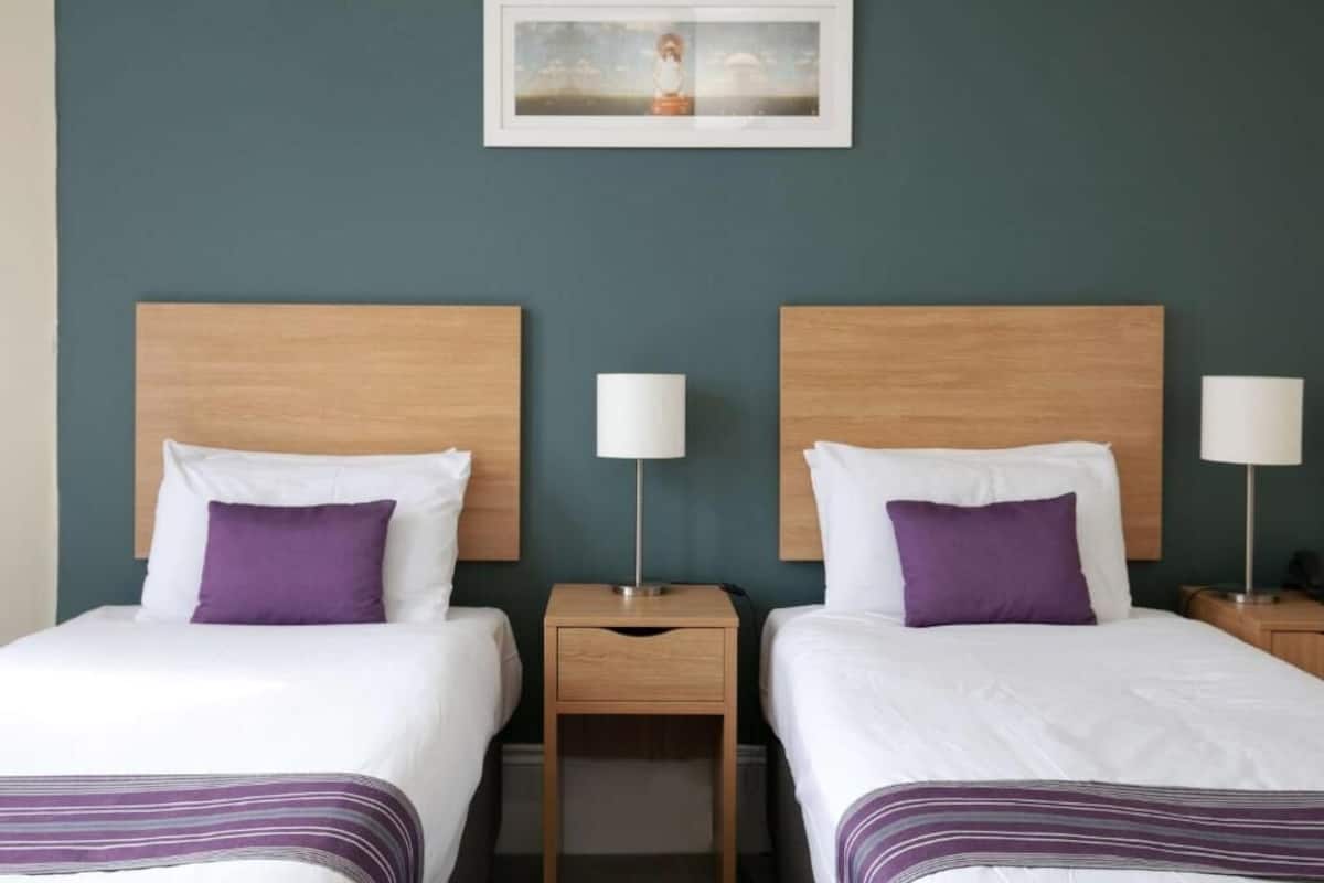 Habitación de hotel con dos camas, cada una con cabecero de madera, ropa de cama blanca y almohadas moradas. Entre las camas hay una mesita de noche de madera con una lámpara a cada lado.