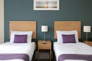 A hotel room with two beds, each with a wooden headboard, white bedding, and purple pillows. There is a small wooden nightstand between the beds with a lamp on each side.