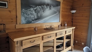 Interior de cabana de madeira com uma grande fotografia emoldurada, uma escrivaninha de madeira com gavetas, uma lâmpada e uma cadeira.
